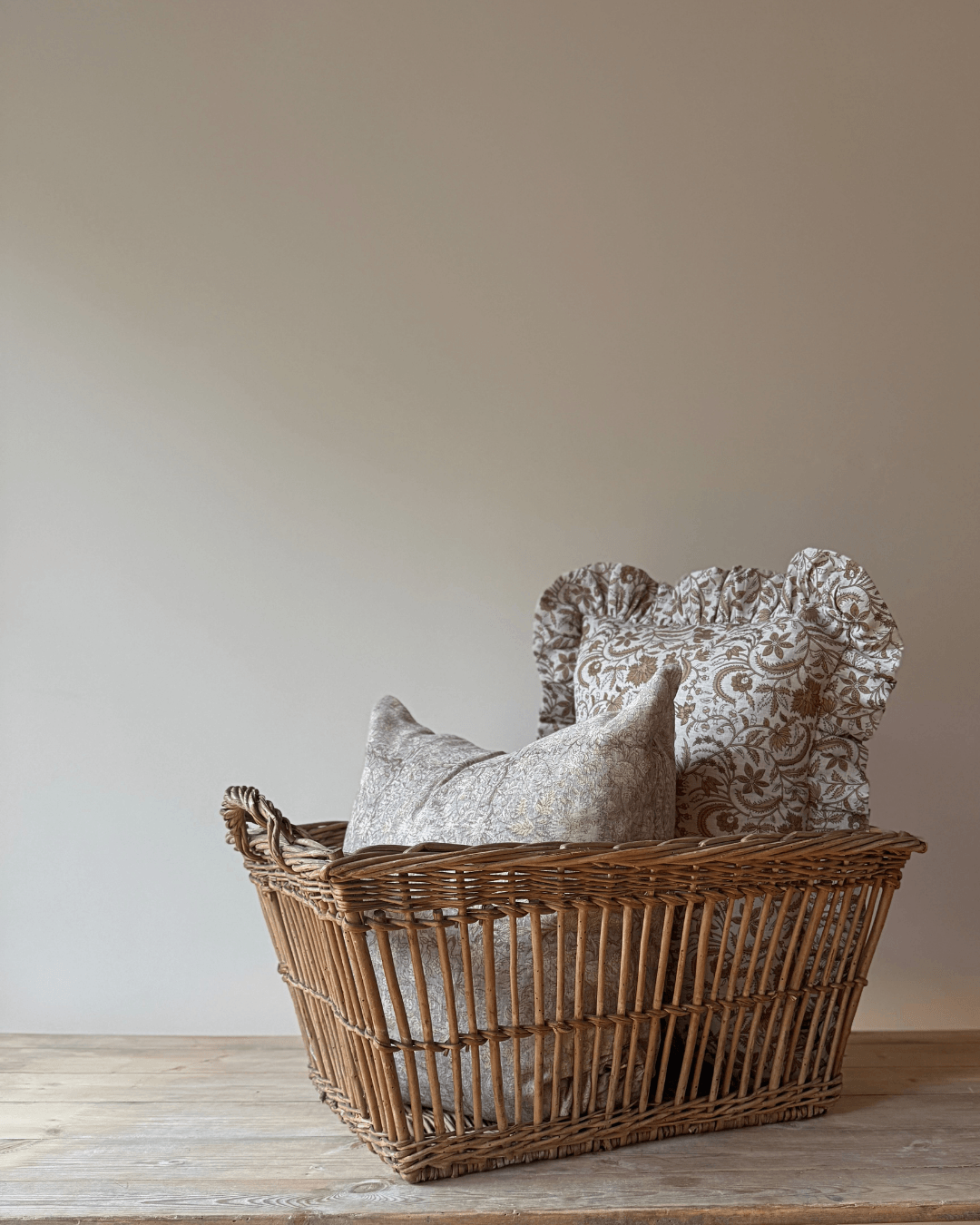 A natural wicker basket filled with two decorative throw pillows, one in a light beige patterned fabric and one in a textured taupe fabric, set on a rustic wooden table against a plain beige wall.