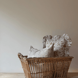 A natural wicker basket filled with two decorative throw pillows, one in a light beige patterned fabric and one in a textured taupe fabric, set on a rustic wooden table against a plain beige wall.