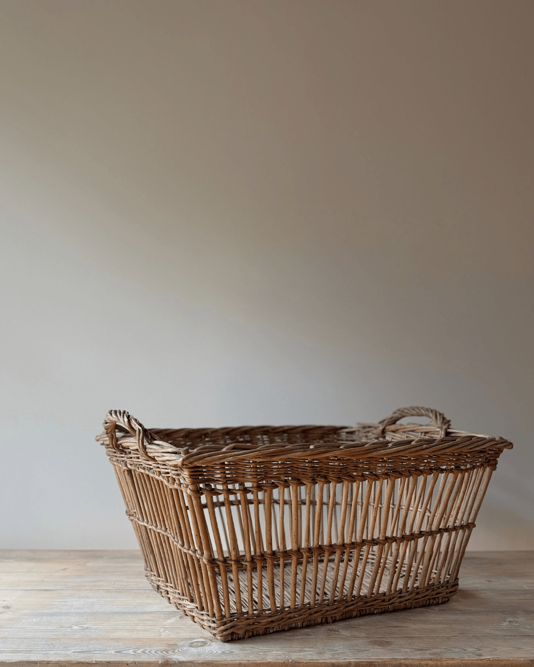 A rustic woven wicker storage basket with integrated handles, resting on a light wooden surface against a neutral beige wall.