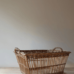 A rustic woven wicker storage basket with integrated handles, resting on a light wooden surface against a neutral beige wall.