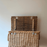 A natural woven antique wicker basket with a hinged lid, shown open on a wooden surface against a plain beige wall.