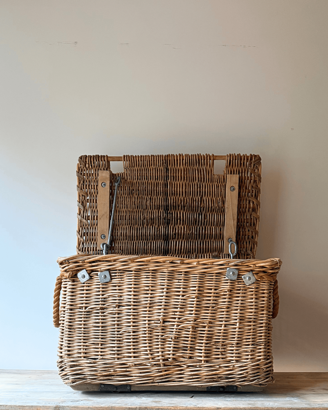 A natural woven antique wicker basket with a hinged lid, shown open on a wooden surface against a plain beige wall.