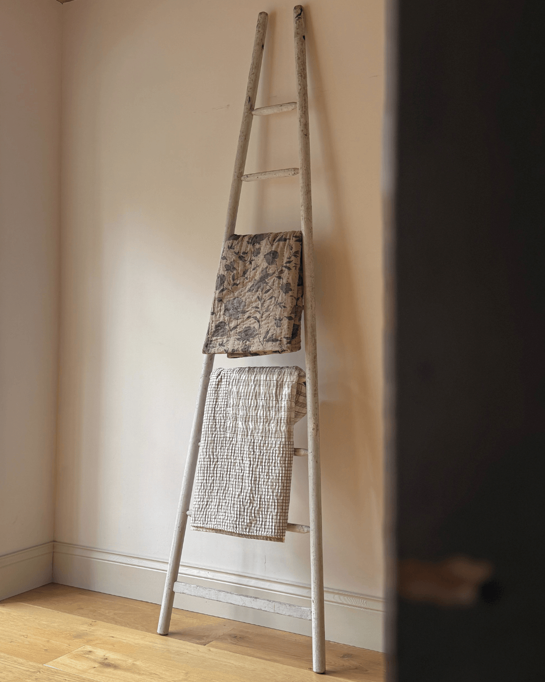 A rustic antique wooden ladder leaning against a cream wall on a light wood floor, with two textured towels draped over its rungs.