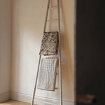A rustic antique wooden ladder leaning against a cream wall on a light wood floor, with two textured towels draped over its rungs.