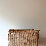 A natural wicker storage basket with a hinged lid and silver metal latches, resting on a light wooden surface against a plain beige wall.