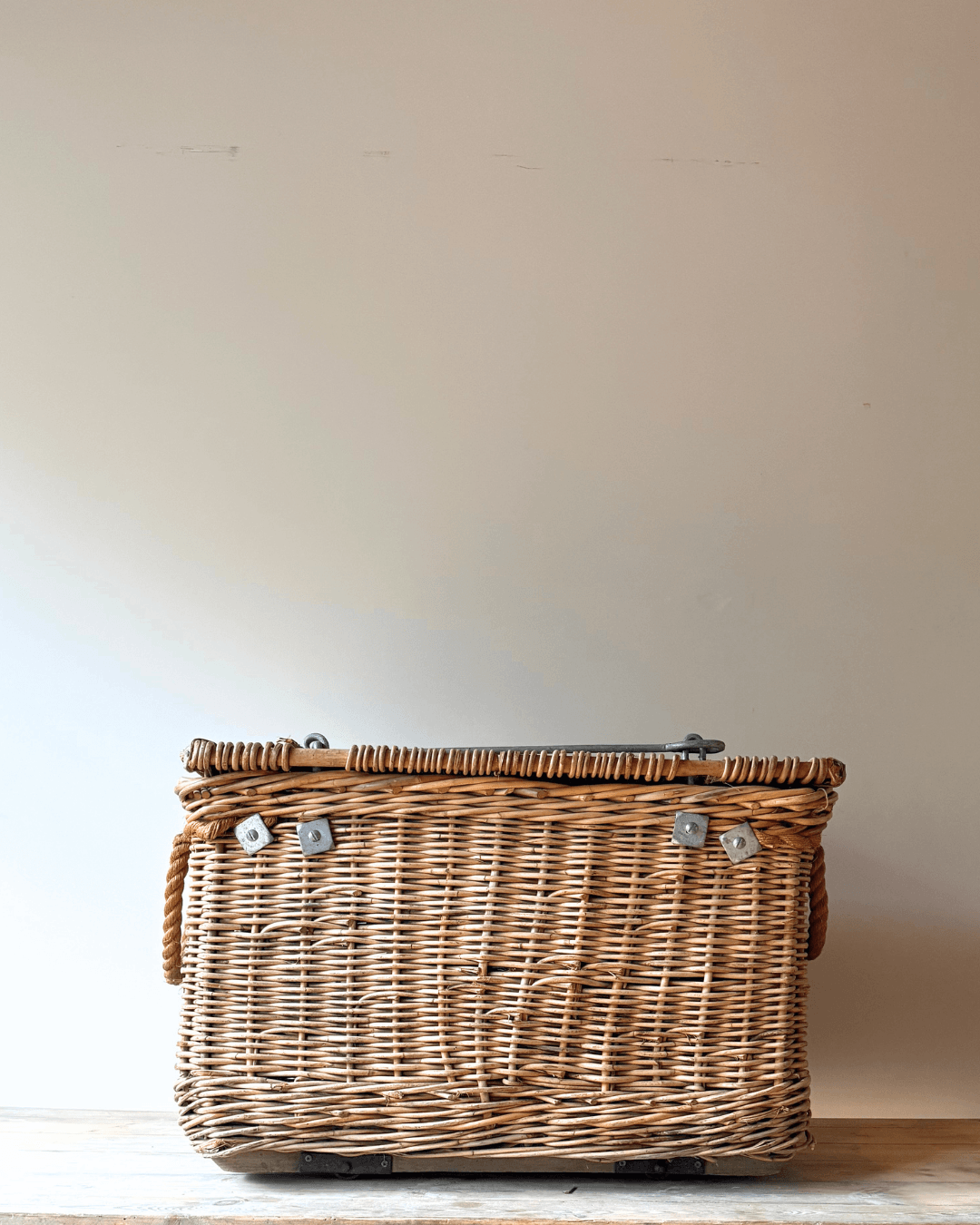 A natural wicker storage basket with a hinged lid and silver metal latches, resting on a light wooden surface against a plain beige wall.