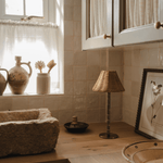 Bamboo table lamp with a wicker shade on a kitchen worktop, with a stone sink and stoneware jugs on the windowsill