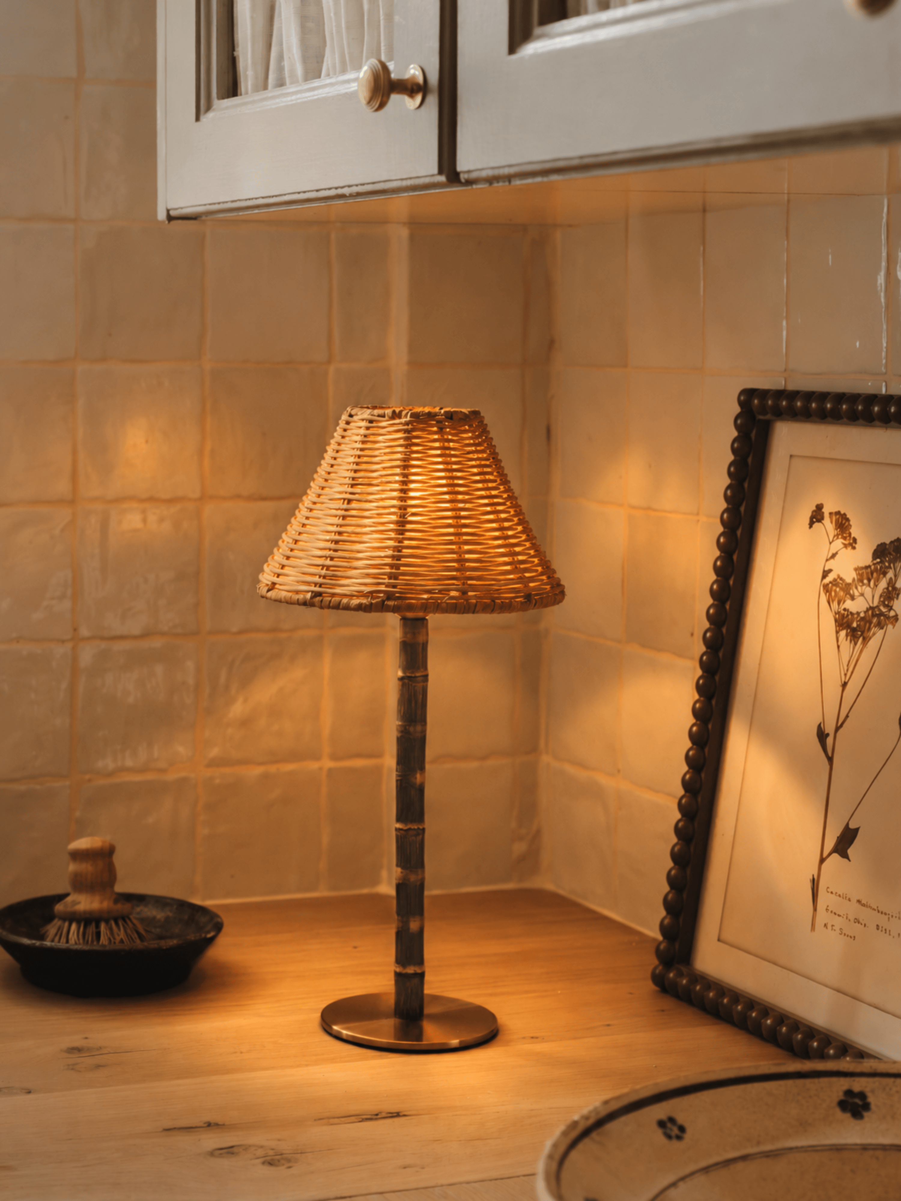 The Raffles cordless lamp glowing warm in a kitchen corner, its wicker shade on a bamboo stem and solid brass base