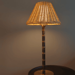 The Raffles lamp lit, casting a soft glow through its woven wicker shade, on a dark round side table