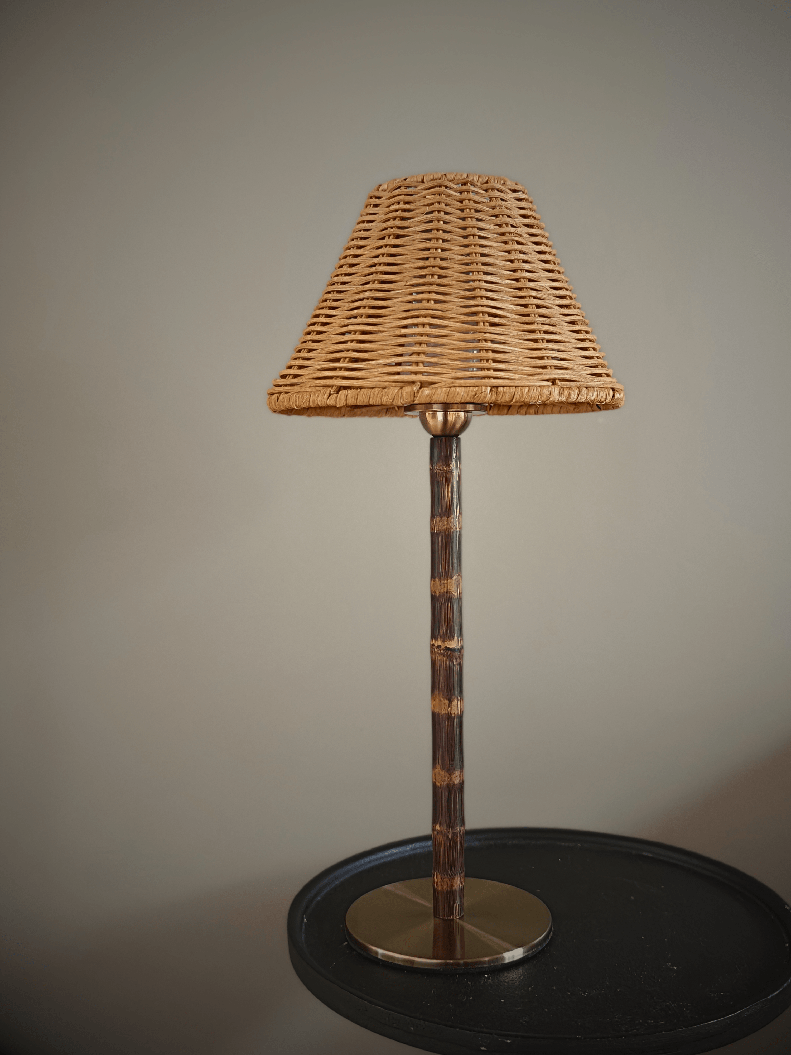 Cordless wicker shade lamp with a bamboo stem and brass base, unlit, on a dark side table against a farrow and ball mouses back wall