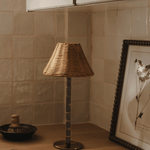 The Raffles bamboo and wicker lamp switched off on a wooden worktop, beside a framed botanical print