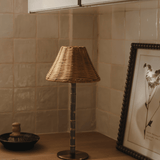 The Raffles bamboo and wicker lamp switched off on a wooden worktop, beside a framed botanical print