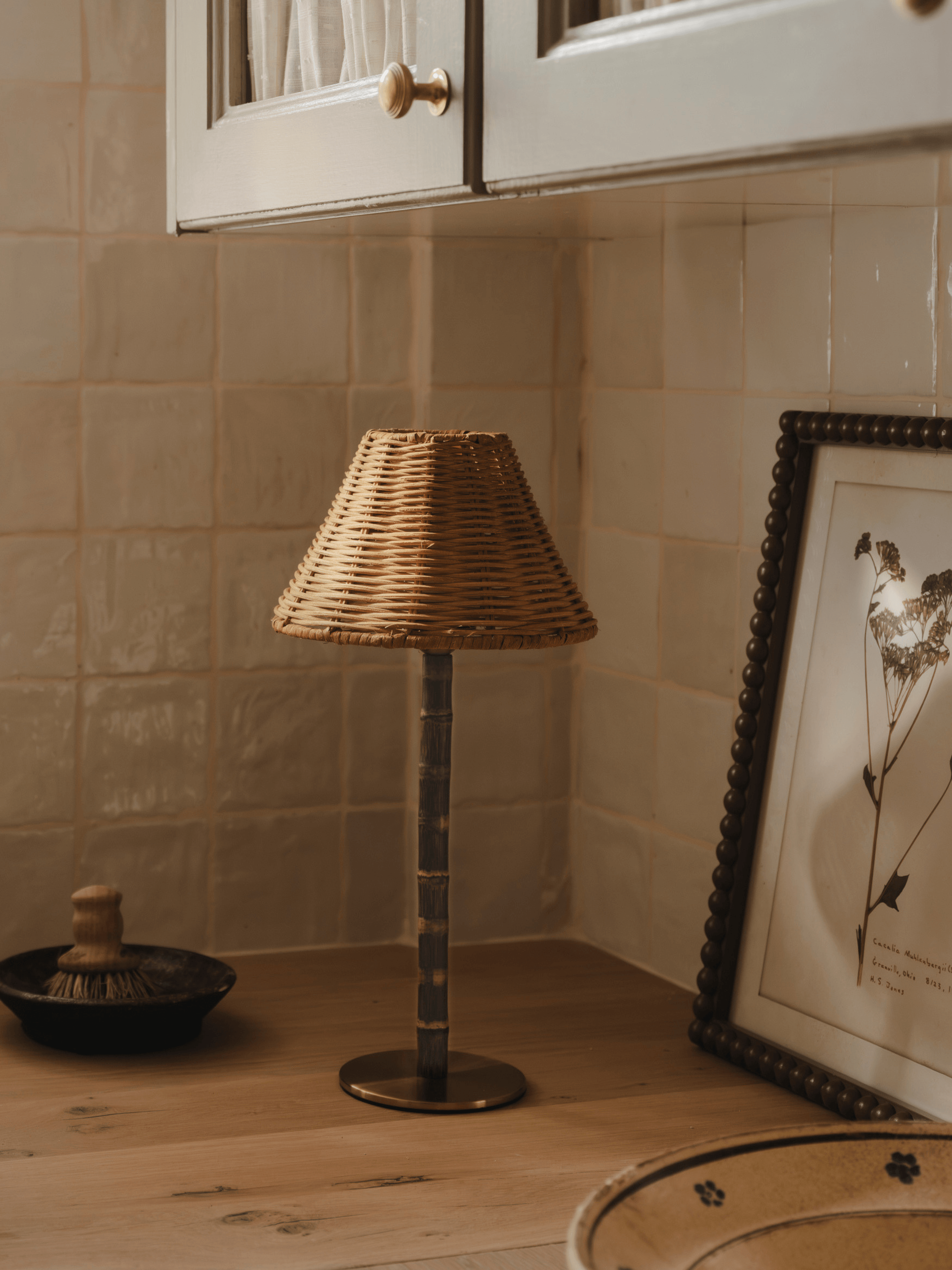 The Raffles bamboo and wicker lamp switched off on a wooden worktop, beside a framed botanical print