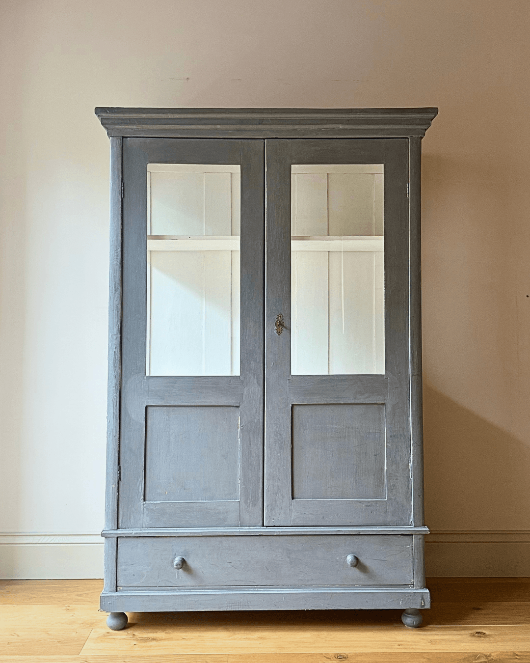 A tall, two-door antique display cabinet or armoire in a distressed blue  finish, featuring glass-paneled doors and a single drawer at the bottom, set against a plain beige wall and light wood floor.
