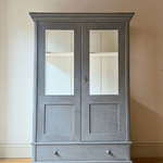 A tall, two-door antique display cabinet or armoire in a distressed blue  finish, featuring glass-paneled doors and a single drawer at the bottom, set against a plain beige wall and light wood floor.
