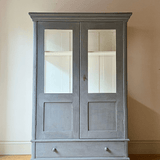 A tall, two-door antique display cabinet or armoire in a distressed blue  finish, featuring glass-paneled doors and a single drawer at the bottom, set against a plain beige wall and light wood floor.