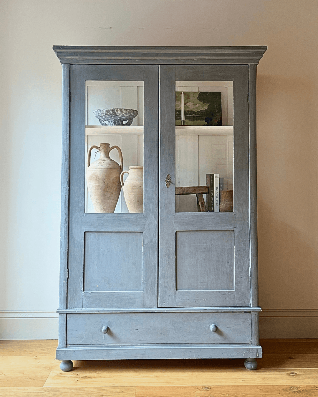 A tall, distressed gray wooden cabinet with two glass-paneled doors and a single drawer at the bottom, styled with vintage ceramic vases on a light wood floor against a neutral beige wall.