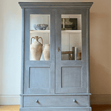 A tall, distressed gray wooden cabinet with two glass-paneled doors and a single drawer at the bottom, styled with vintage ceramic vases on a light wood floor against a neutral beige wall.