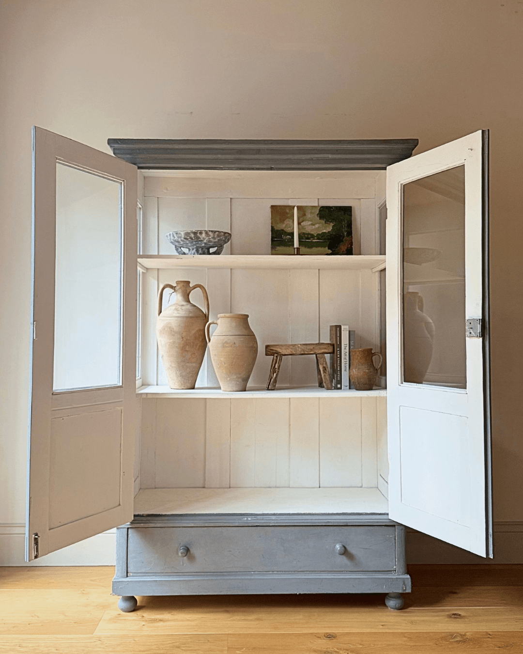 A tall blue antique display cabinet for sale with two open doors and a blue painted base and top, styled with rustic vases and a small wooden stool on a light wood floor against a beige wall.