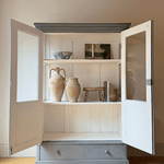 A tall blue antique display cabinet for sale with two open doors and a blue painted base and top, styled with rustic vases and a small wooden stool on a light wood floor against a beige wall.