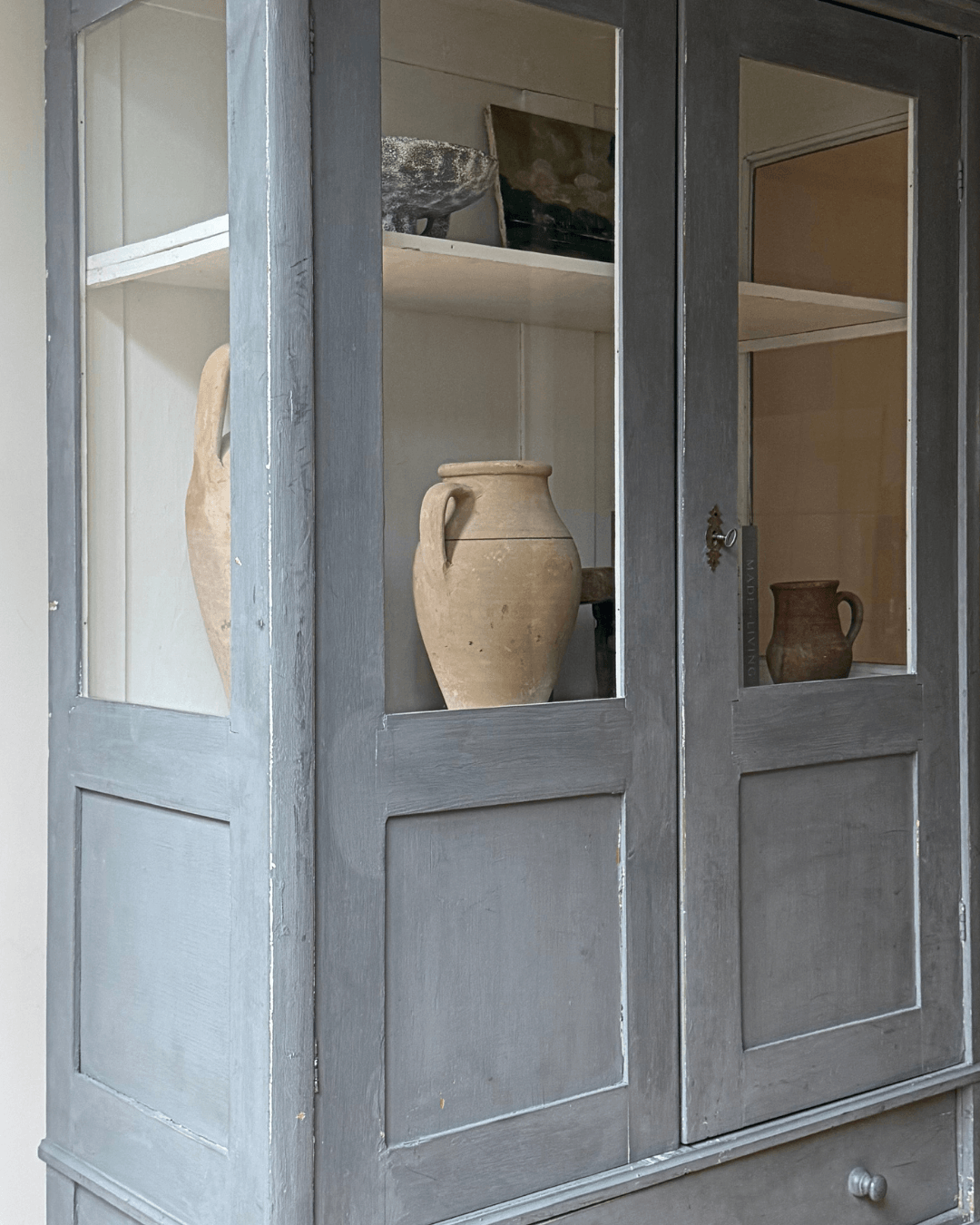 Blue antique wooden cabinet with glass doors, displaying vintage ceramic vases on its shelves against a light background.
