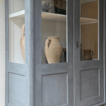 Blue antique wooden cabinet with glass doors, displaying vintage ceramic vases on its shelves against a light background.