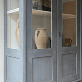 Blue antique wooden cabinet with glass doors, displaying vintage ceramic vases on its shelves against a light background.