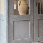 A close-up of an antique wooden cabinet or door with a small window showing a beige ceramic vase inside.