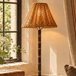 The Raffles bamboo rechargeable lamp switched on at a cottage window, its wicker shade glowing warm over a bamboo stem and brass base